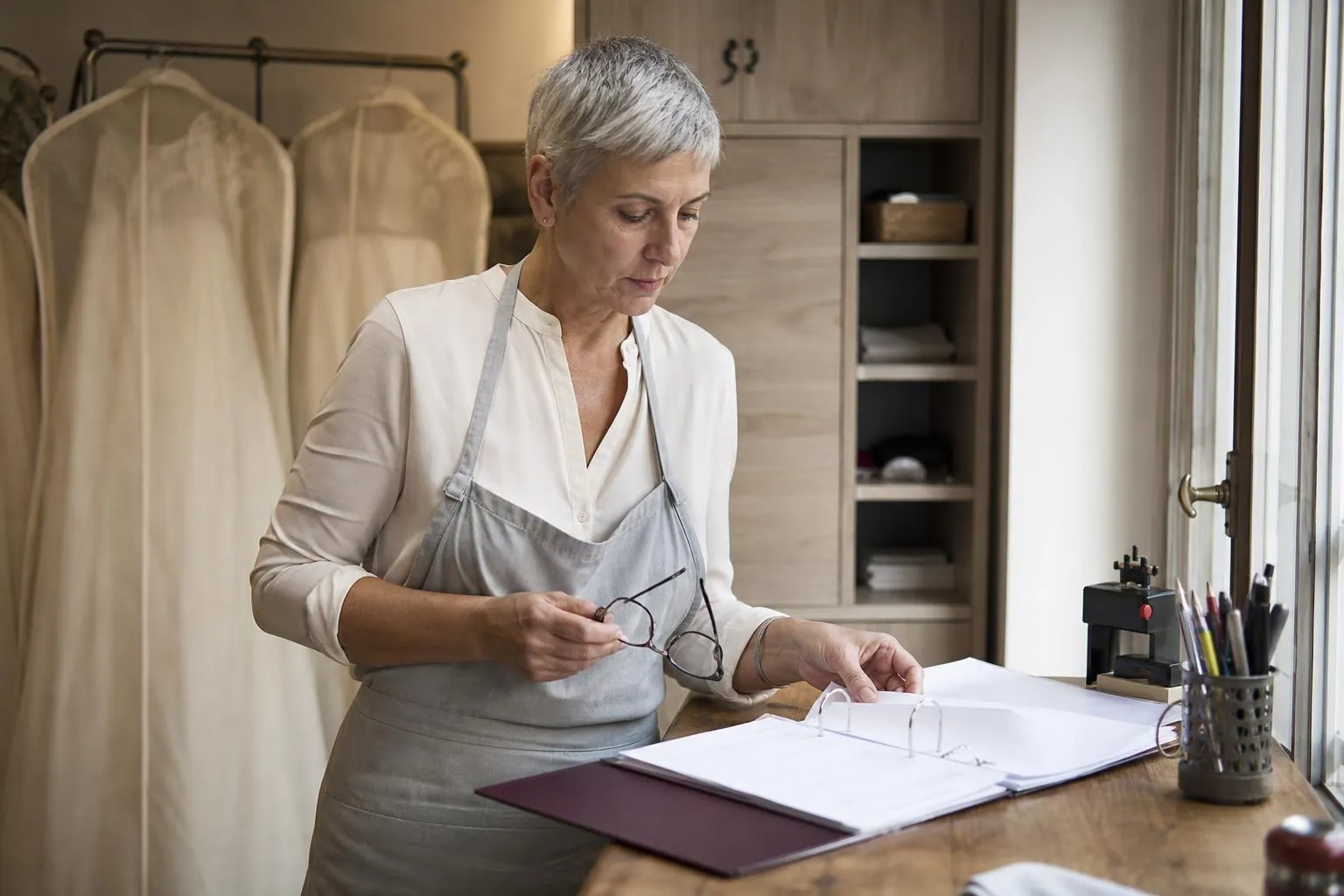 Couturière d’une cinquantaine d’années debout à son petit bureau d’atelier, feuilletant un classeur de devis et de conditions de vente près d’une fenêtre claire