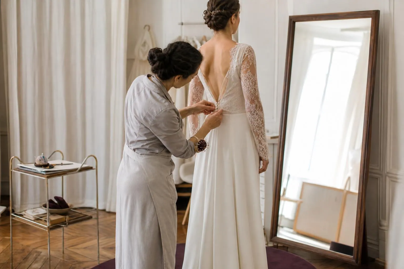Créatrice agenouillée derrière une cliente en robe de mariée sur un podium bas, épinglant l’ouverture du dos devant un grand miroir sur pied